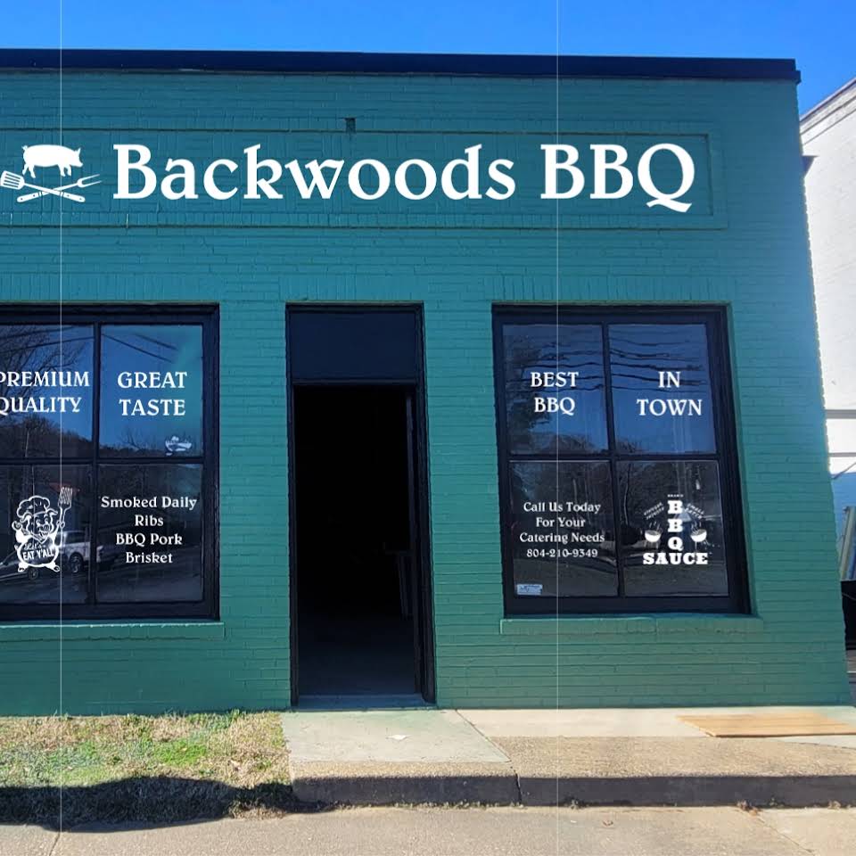 Storefront of Backwoods BBQ, located next to Ace Hardware, vibrant green brick building.