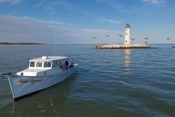 A white boat sitting in calm waters near a tall white lighthouse on a rock island