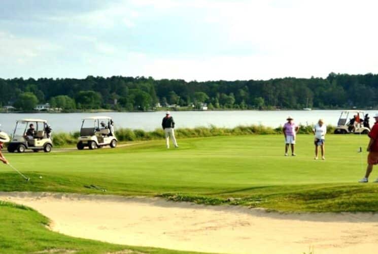 Several golfers and carts on a green with a sand trap by a large lake surrounded by trees