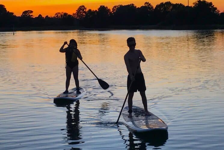 Two people padding stand up paddle boards on a calm lake surrounded by trees at sunset