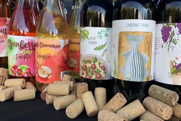 Several bottles of wine with colorful labels lined up on a table with corks