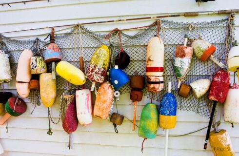 Collage of an old fishing net, fishing pole, and colorful buoys hanging on the side of an exterior wall