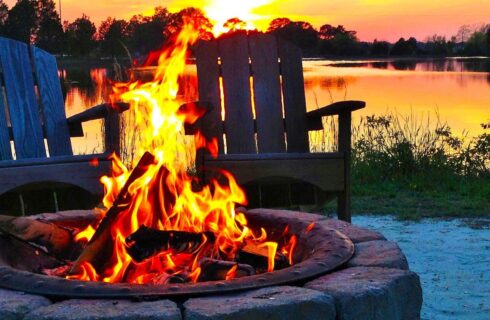 Close up view of lighted fire pit with two Adirondack chairs and calm water in the background