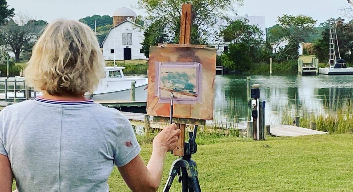 Woman standing in grass painting a picture of white boat docked on the water