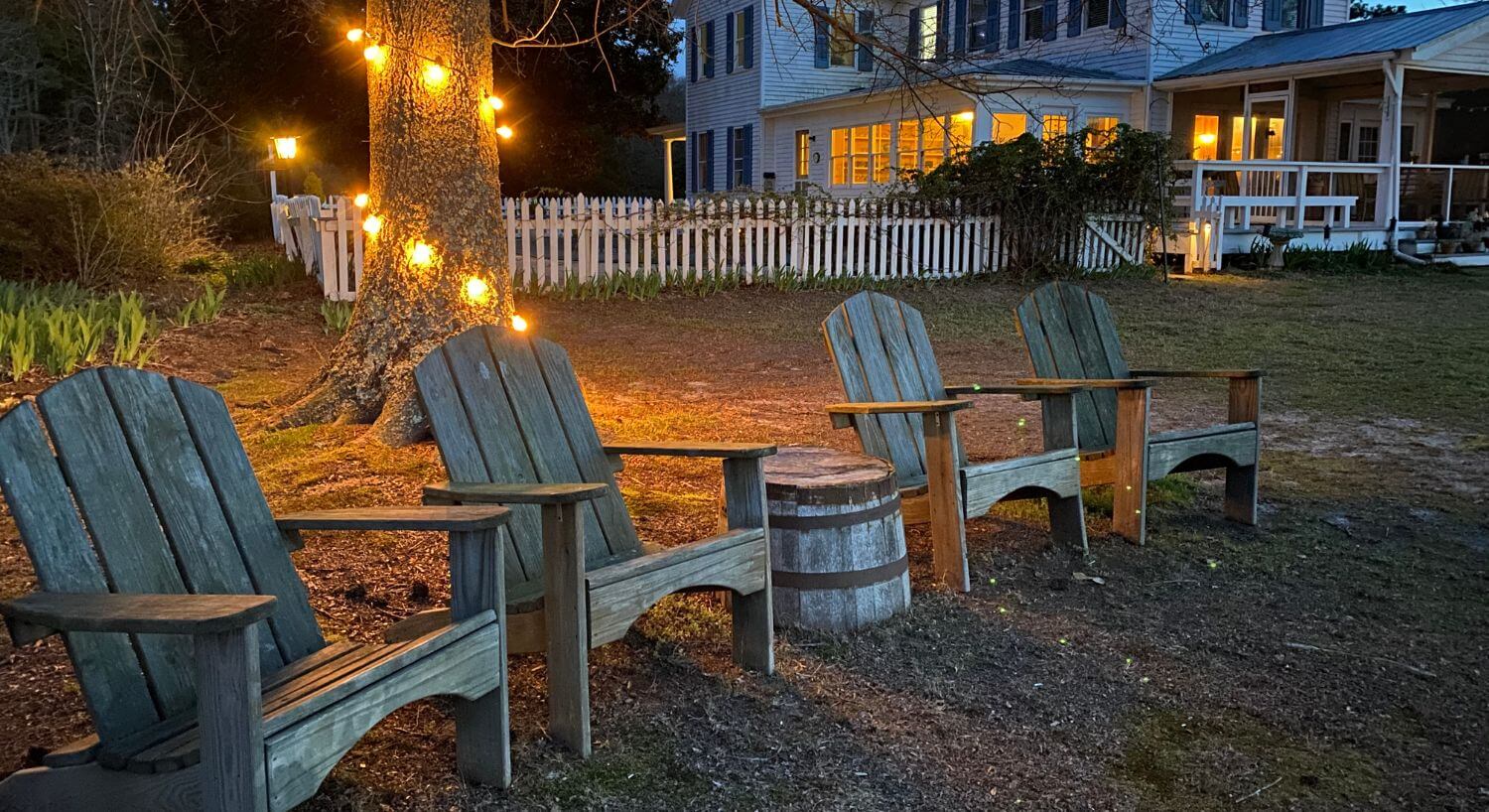 Four wooden Adirondack chairs lined up in the backyard with string lights around the base of a tree trunk at dusk