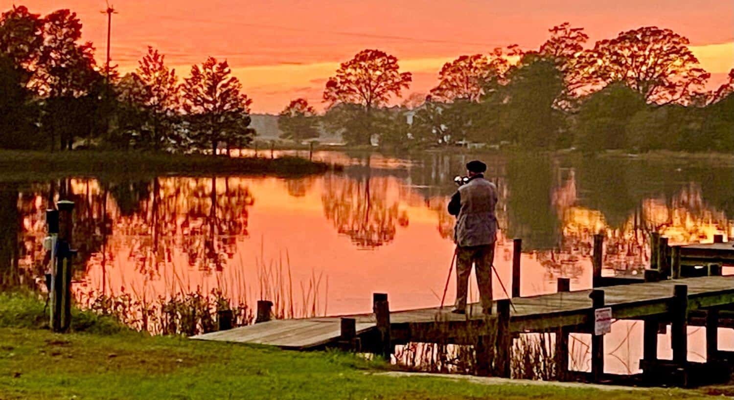 Man with a tripod standing on a pier looking out at calm water at dusk