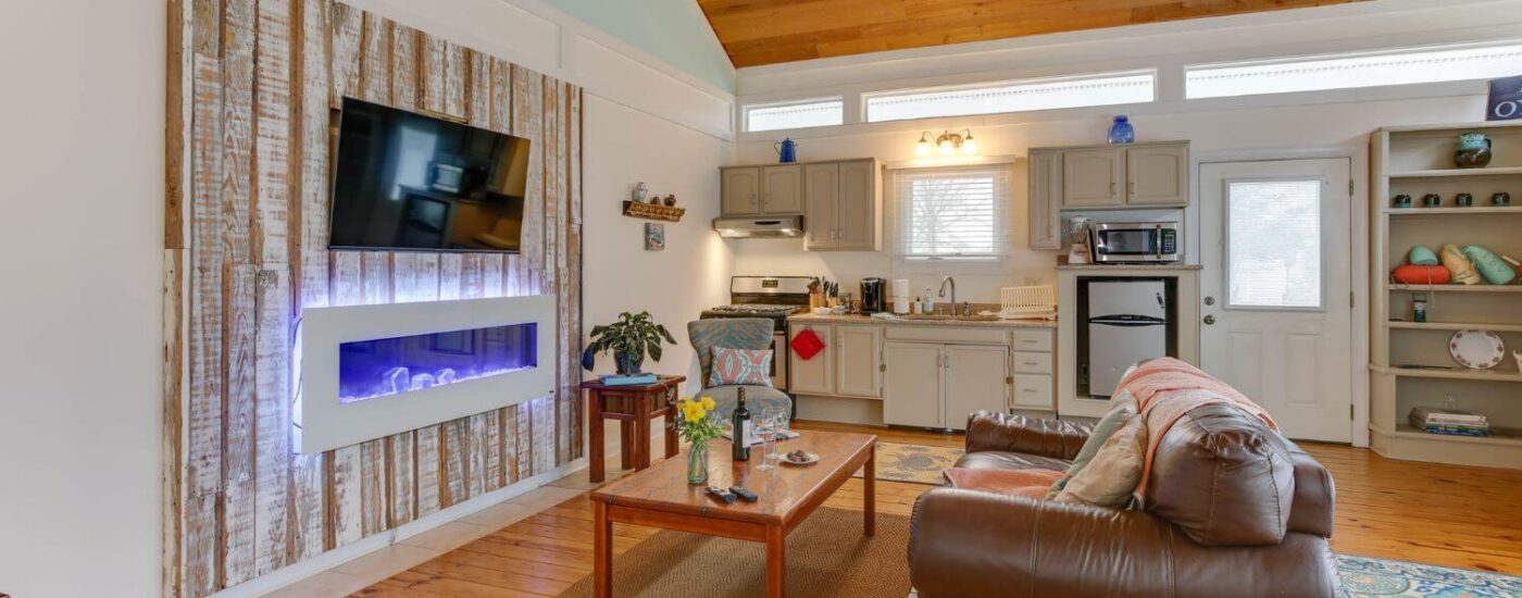 Adjoining living room to bedroom with white walls, hardwood flooring, brown leather couch, wall mounted flat-screen TV, and small kitchen