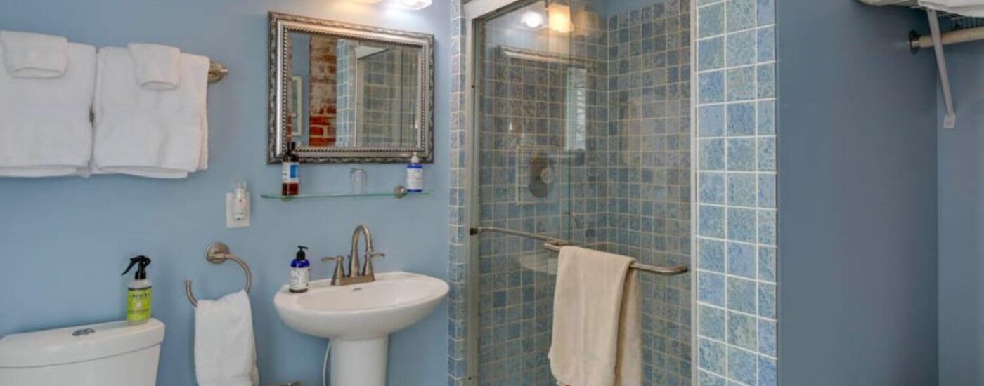 Bathroom with blue walls, light-blue tiled walk-in shower with glass door, white pedestal sink, and silver framed mirror