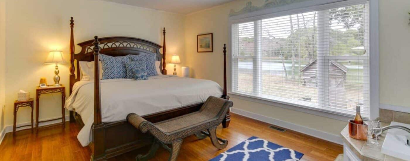 Bedroom with light yellow walls, white trim, hardwood flooring, dark wooden four poster bed, and view to the backyard from a large window