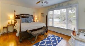 Bedroom with light yellow walls, white trim, hardwood flooring, dark wooden four poster bed, and view to the backyard from a large window