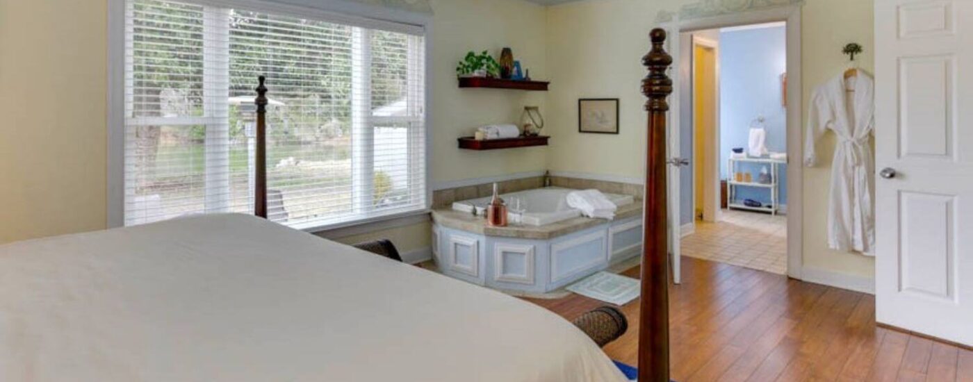 Bedroom with light yellow walls, white trim, hardwood flooring, dark wooden four poster bed, and soaker tub