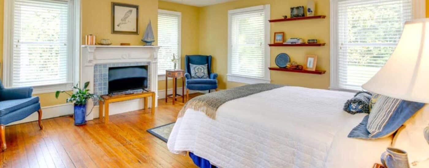 Bedroom with yellow walls, white trim, hardwood flooring, white bedding, two blue upholstered vintage armchairs, and flat-screen TV on small table