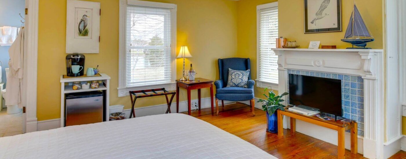 Bedroom with yellow walls, white trim, hardwood flooring, white bedding, blue upholstered vintage armchair, and flat-screen TV on small table