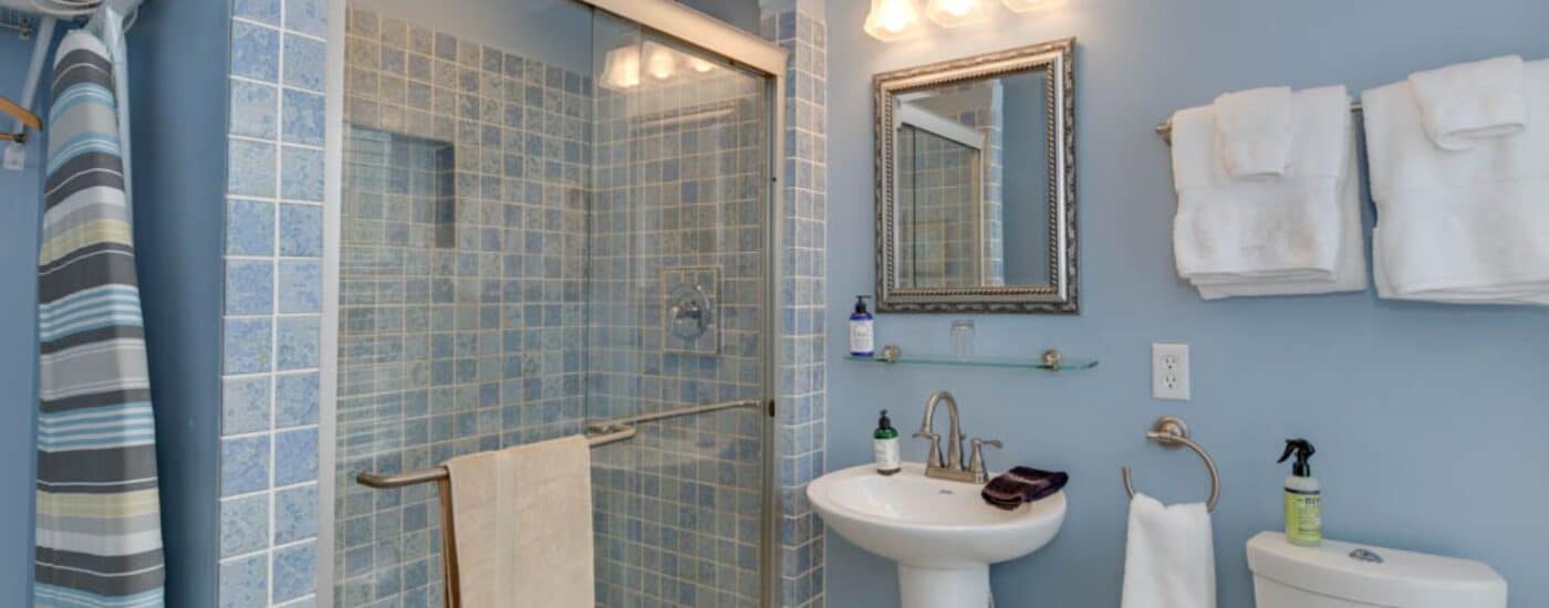 Bathroom with blue walls, light-blue tiled walk-in shower with glass door, white pedestal sink, and silver framed mirror