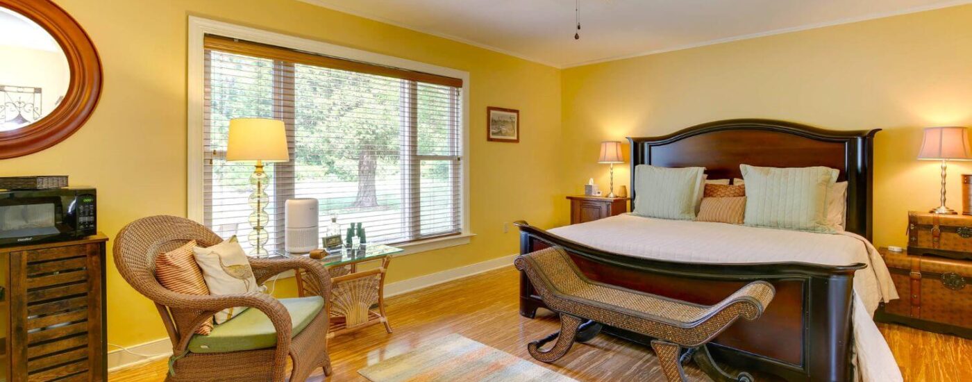 Bedroom with yellow walls, white trim, hardwood flooring, dark wooden bed, and light brown wicker chair with lime green cushion