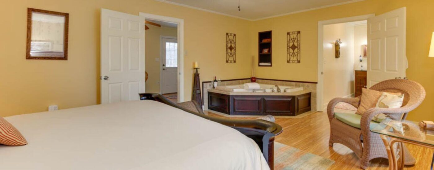 Bedroom with yellow walls, white trim, hardwood flooring, dark wooden bed, light brown wicker chair with lime green cushion, and soaker tub in the corner of the room