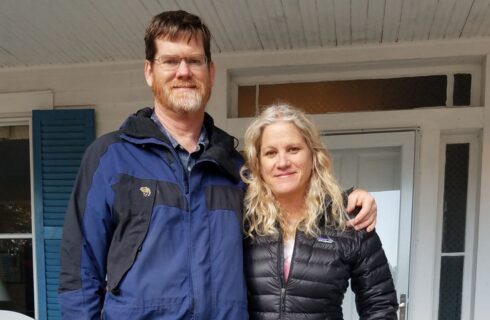 Man and woman in jackets with arms around each other standing on front porch smiling