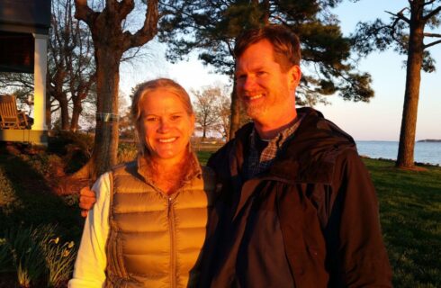 Woman wearing a vest and man wearing a jacket with arms around each other standing in grass