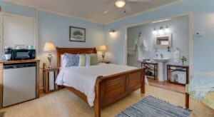 Bedroom with light blue walls, white trim, light colored flooring, wooden bed, mini fridge, and view into bathroom