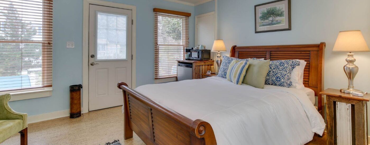 Bedroom with light blue walls, white trim, light colored flooring, wooden bed, mini fridge, and green upholstered vintage armchair