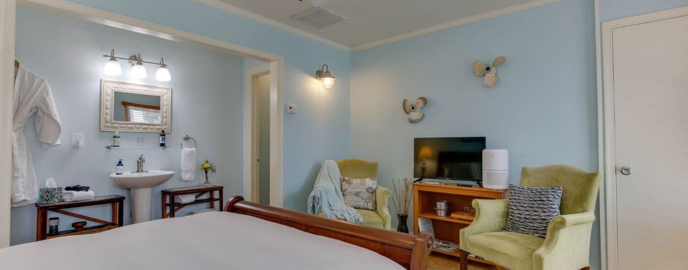 Bedroom with light blue walls, white trim, light colored flooring, wooden bed, sitting area, and view into bathroom