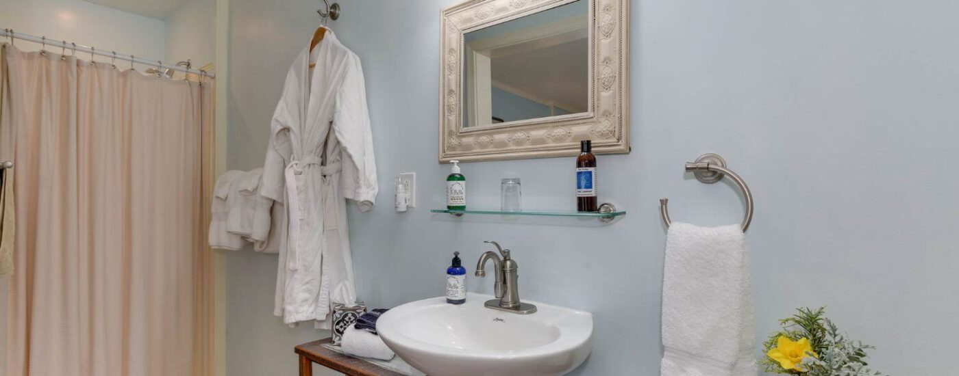 Bathroom with light blue walls, white pedestal sink, framed mirror, and walk-in shower with pale pink curtain