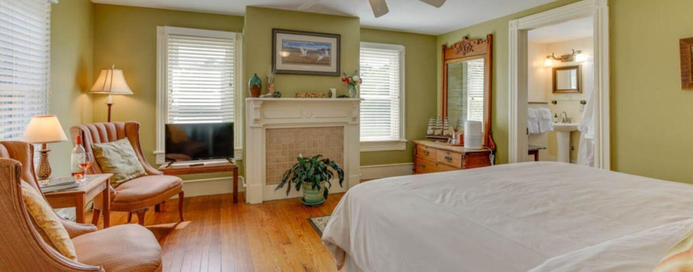 Bedroom with light green walls, white trim, hardwood flooring, white bedding, sitting area, and view into bathroom