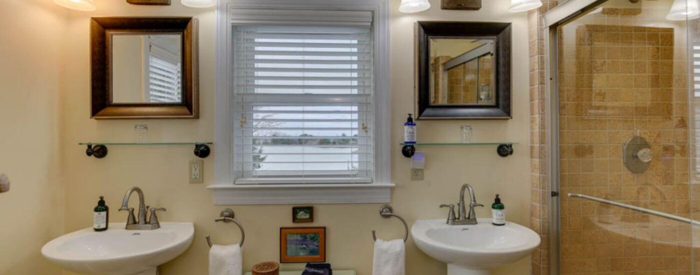 Bathroom with cream walls, two separate white pedestal sinks, and walk-in tan-tiled shower with glass door