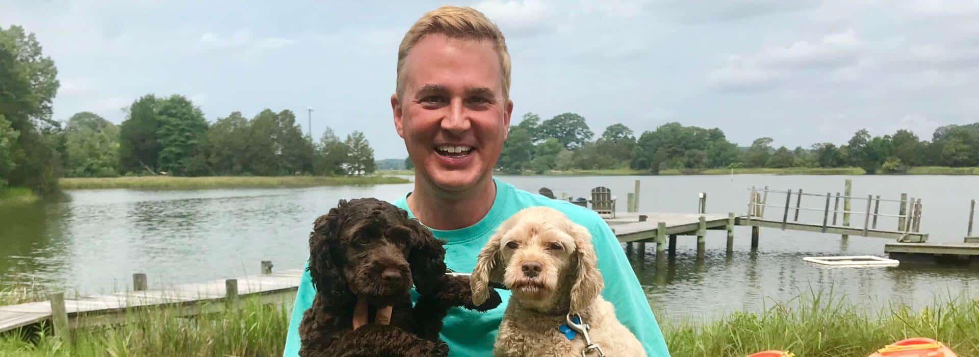 Man holding two dogs smiling for the camera with dock and water in the background