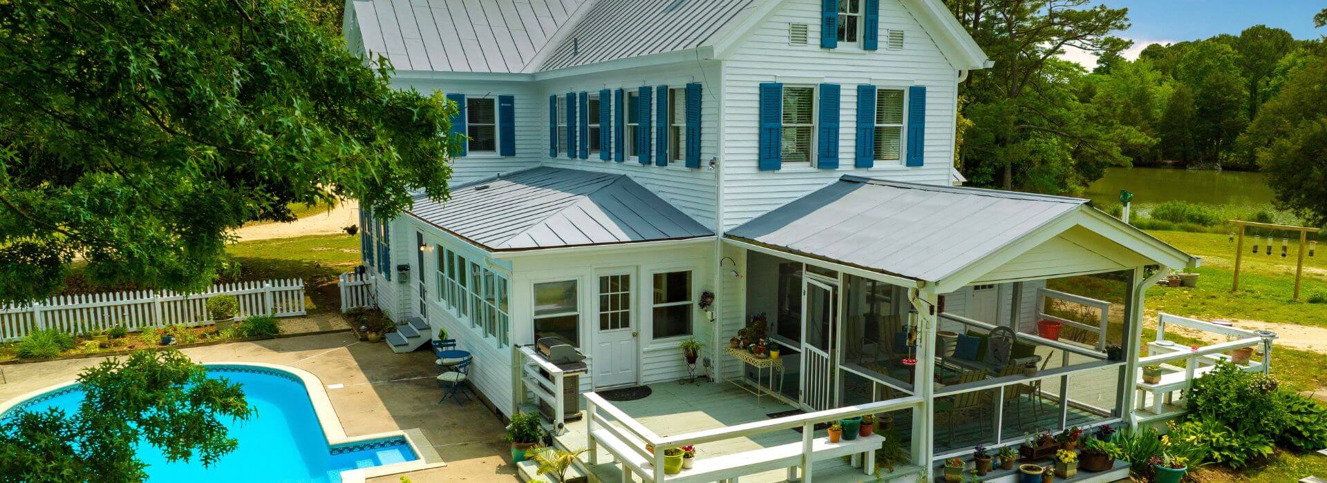 Exterior view of property painted white with blue shutters and a pool surrounded by green trees