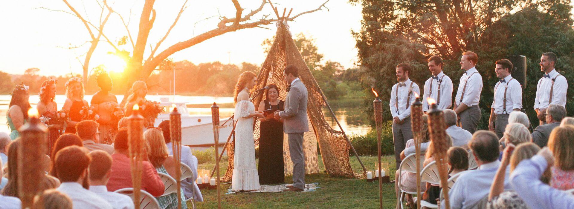 Bride and groom standing in front of family and friends at a wedding on the lawn with the small white boat on the water in the background