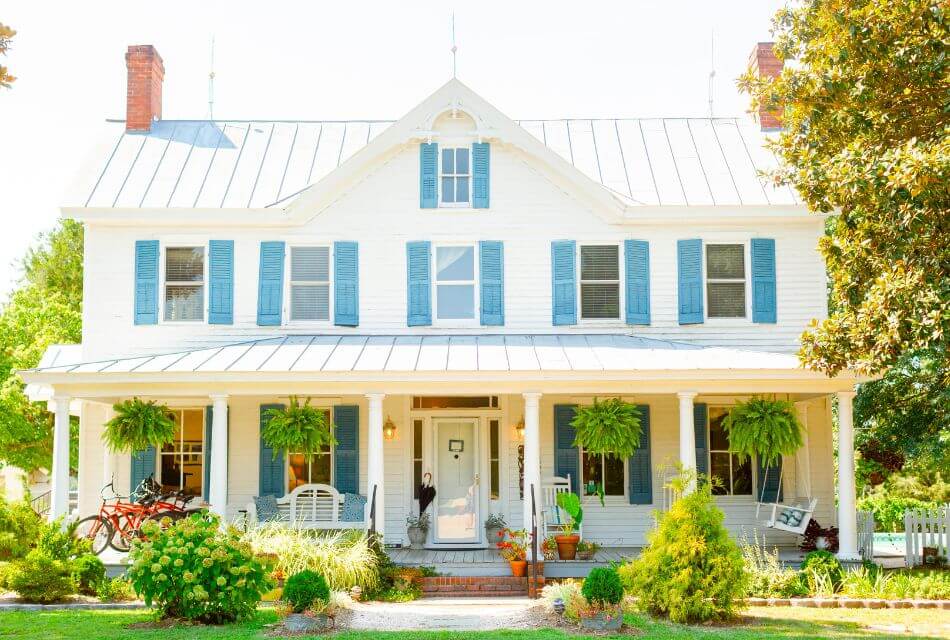 Exterior view of property painted white with teal shutters and front porch with white bench, white rocking chairs, and white bench swing