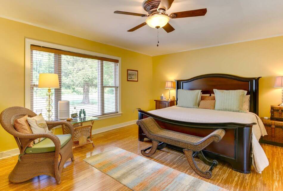 Bedroom with yellow walls, white trim, hardwood flooring, dark wooden bed, and light brown wicker chair with lime green cushion