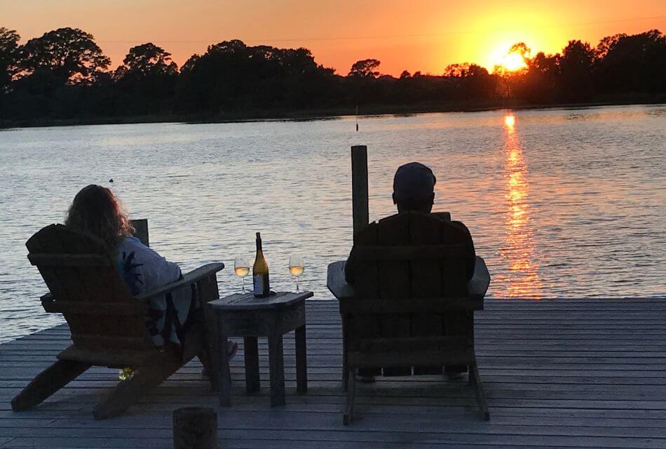 Two people sitting on a dock facing the setting sun in the background