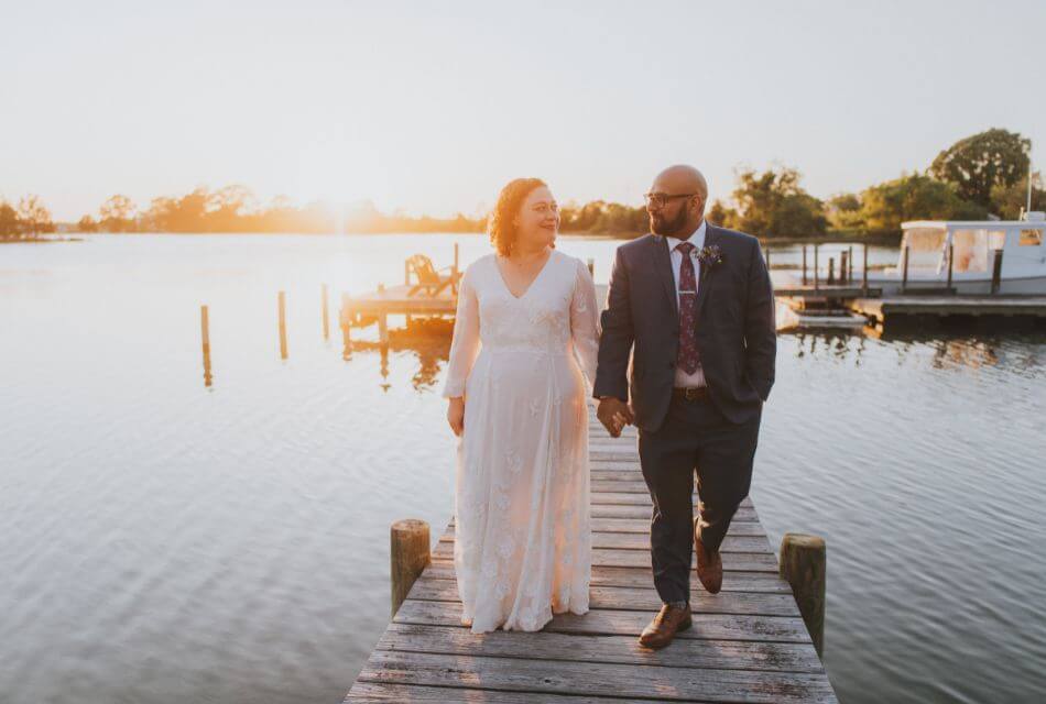 Woman in white dress and man in dark suit walking on a pier surrounded by water with sun in the background