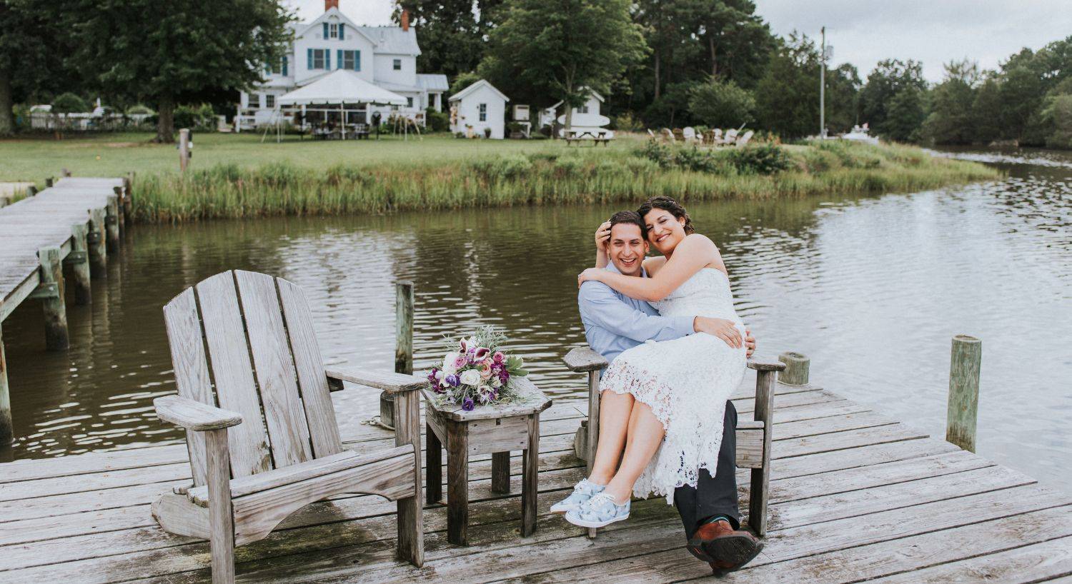 Bride sitting on groom's lap in Adirondack chair on dock with water and property in the background