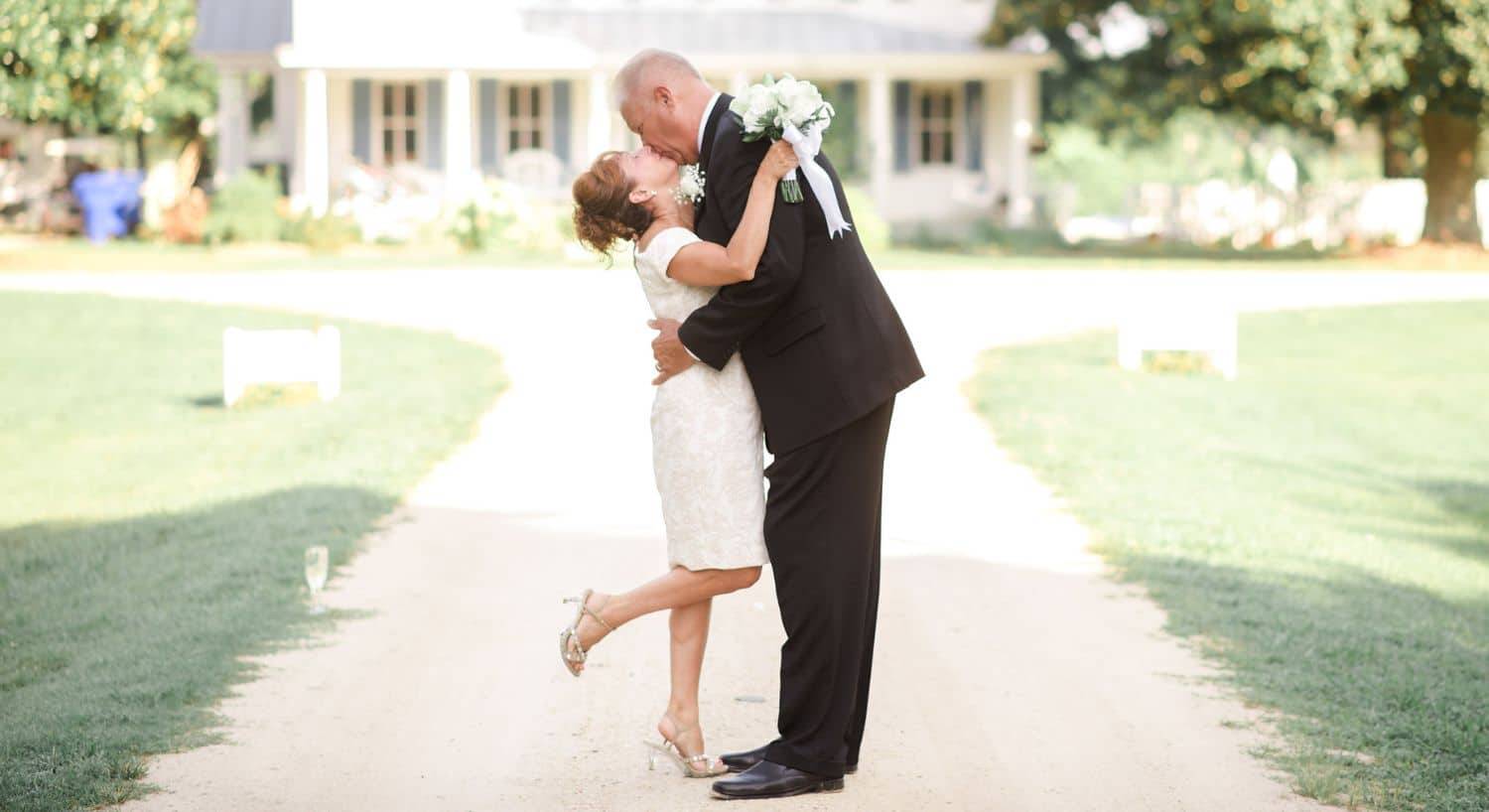 Bride and groom standing and kissing on a gravel path with property in the background