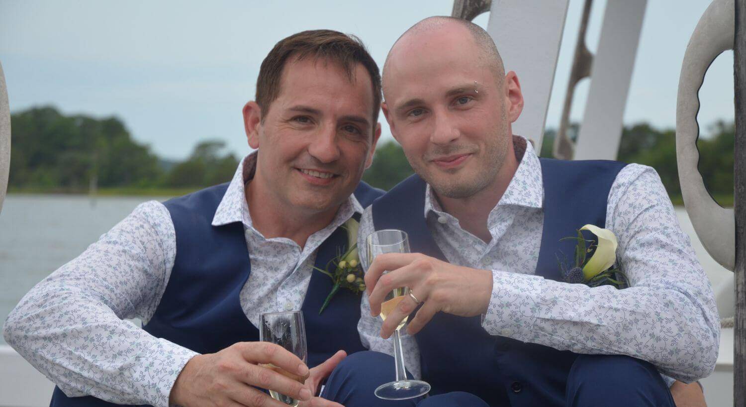 Two men wearing navy vests holding Champagne flutes smiling at the camera