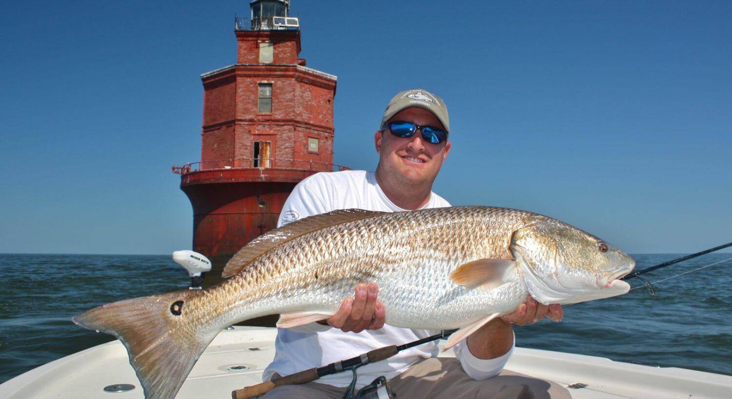 Man holding large gold and white fish on a boat with red brick lighthouse in the background