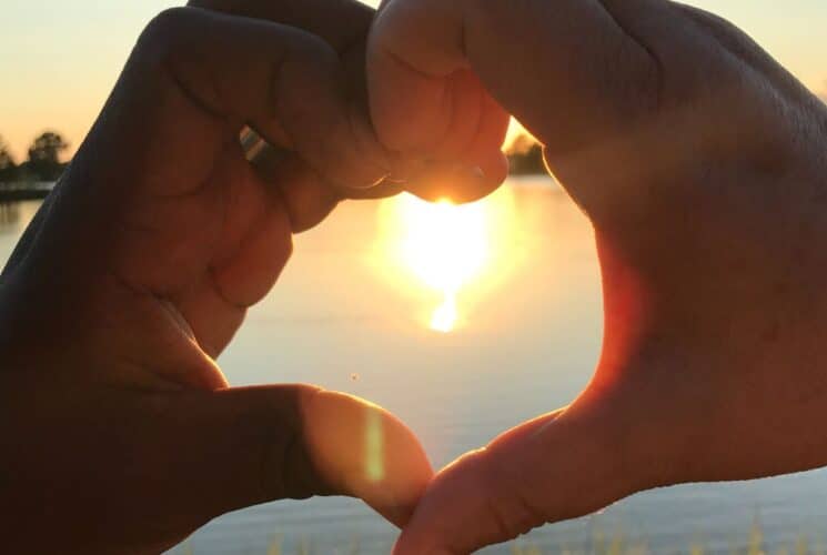 Person's fingers in a shape of a heart with reflecting sun on the water in the middle of the heart