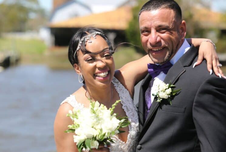 Bride and groom with arms around each other smiling at the camera with water in the background