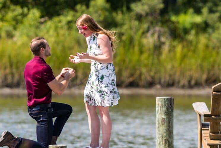 Man down on one knee while woman smiles enthusiastically at man at the end of a doc with calm water in the background