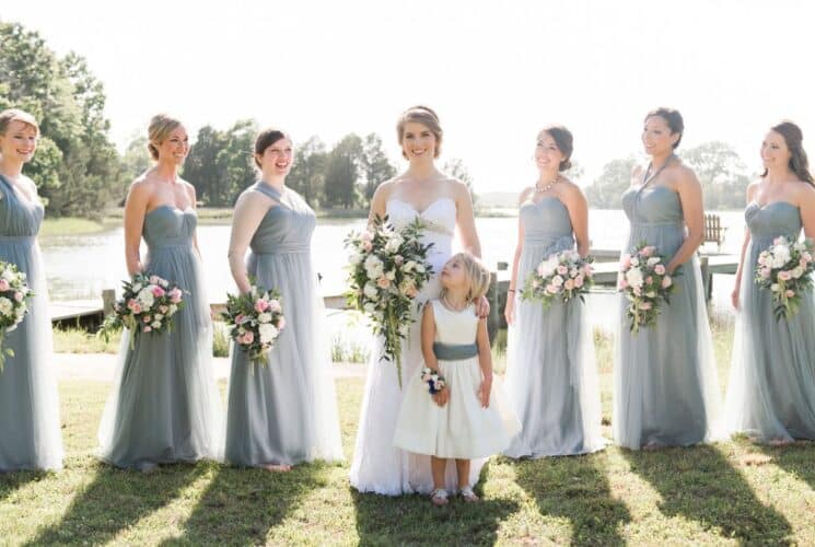 Bride standing with flower girl and bridesmaids standing on grass with dock and water in the background